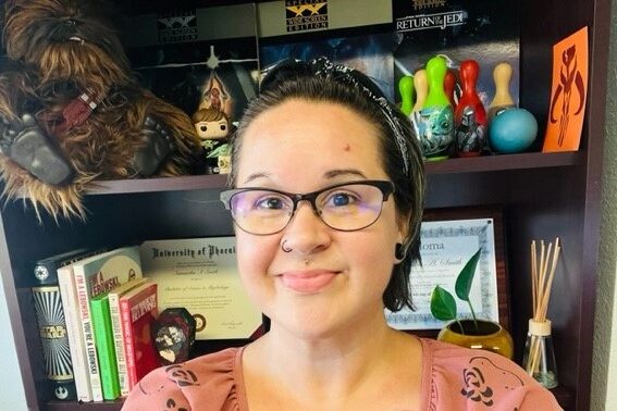 Woman with glasses in a bookstore or office setting, smiling at the camera.