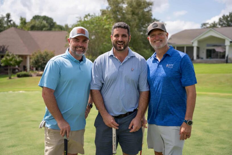 Michael Hobbs, Stelios Peterson, and Corey O'Kelly of Team Centennial Bank pose together on a golf course.